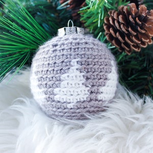 May include: A spherical Christmas ornament covered in a light gray crocheted yarn casing featuring a white stitched pine tree pattern. The ornament is topped with a silver-colored metal cap and a small wire loop for hanging purposes.