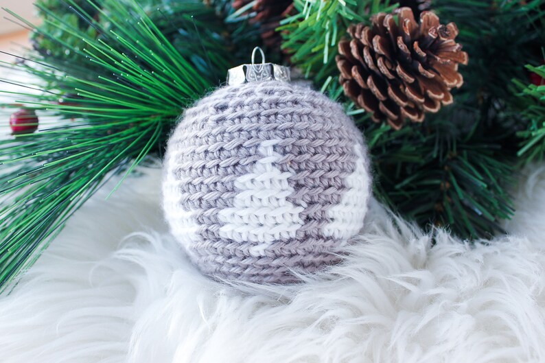 May include: A spherical Christmas ornament covered in a light gray crocheted yarn casing featuring a white stitched pine tree pattern. The ornament is topped with a silver-colored metal cap and a small wire loop for hanging purposes.