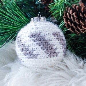 May include: A spherical Christmas ornament covered in white and light gray crocheted yarn with a textured, fuzzy appearance. The ornament features a silver-colored metal cap at the top with a small loop for hanging, showcasing a detailed pattern.