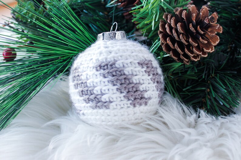 May include: A spherical Christmas ornament covered in white and light gray crocheted yarn with a textured, fuzzy appearance. The ornament features a silver-colored metal cap at the top with a small loop for hanging, showcasing a detailed pattern.