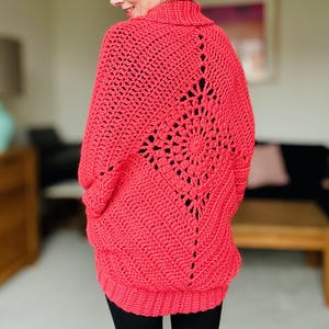 May include: A bright pink crocheted cardigan with a large openwork design on the back. The cardigan has a relaxed fit and a high neckline.