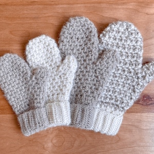 May include: Three crocheted mittens in beige and white. The mittens are made with a textured stitch and have a simple cuff.