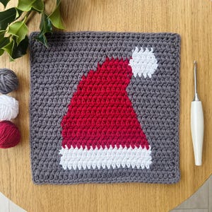 May include: A gray crochet square with a red and white Santa hat design in the center. The hat has a white pom-pom on top and a white trim around the brim.