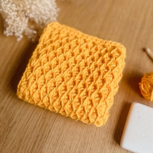 May include: A yellow crocheted square with a textured pattern. The square is lying on a wooden surface.