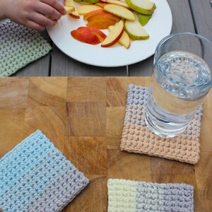 May include: Four crocheted coasters in different color combinations. The coasters are made with a textured stitch and are in shades of blue, gray, beige, and yellow. The coasters are on a wooden surface with a glass of water and a plate of fruit.