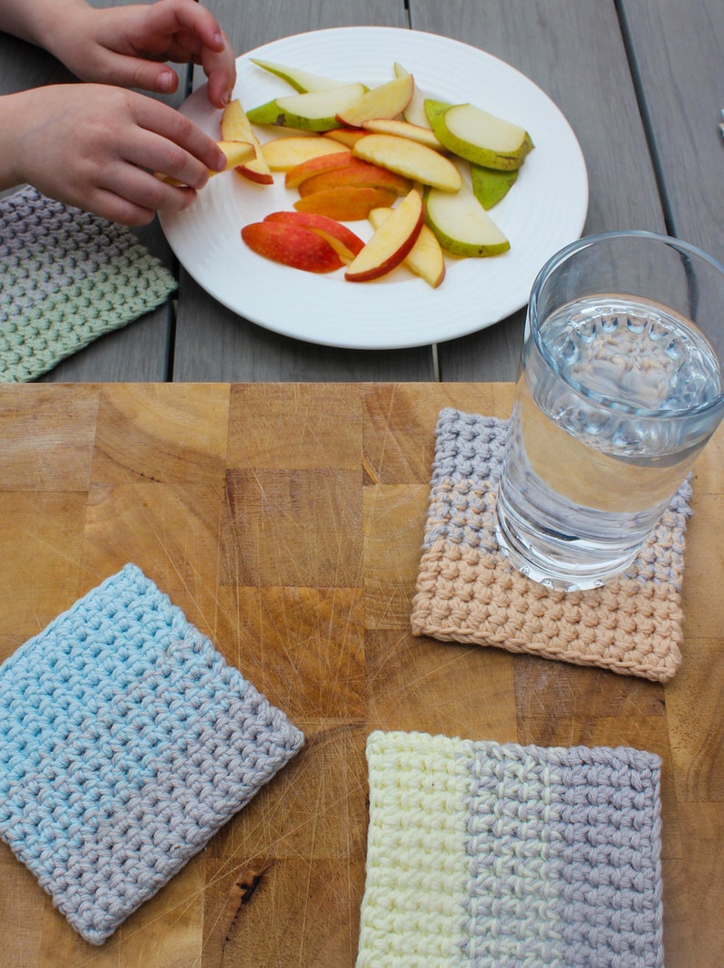 May include: Four crocheted coasters in different color combinations. The coasters are made with a textured stitch and are in shades of blue, gray, beige, and yellow. The coasters are on a wooden surface with a glass of water and a plate of fruit.
