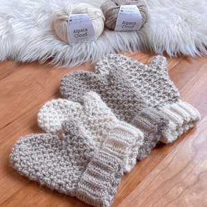 May include: Three pairs of crocheted mittens in beige and white. The mittens are made with a textured stitch and have a ribbed cuff. The yarn balls are labeled "Alpaca Cloud" in white and beige.