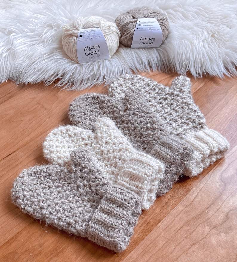 May include: Three pairs of crocheted mittens in beige and white. The mittens are made with a textured stitch and have a ribbed cuff. The yarn balls are labeled "Alpaca Cloud" in white and beige.