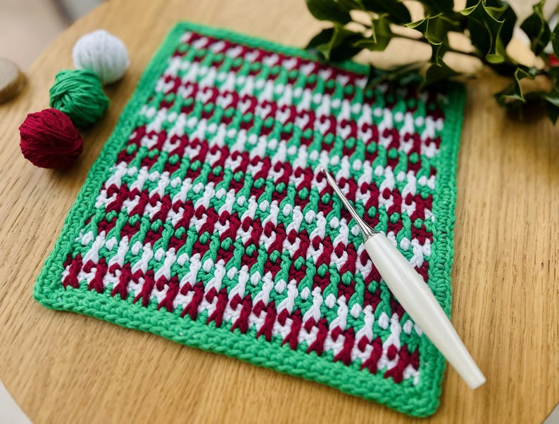May include: A green, white, and red crocheted square with a white crochet hook lying on top. The square is made with a repeating pattern of interlocking loops.