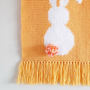 May include: A crocheted wall hanging with a white bunny silhouette and a pink and orange pom-pom. The hanging has a yellow fringe.