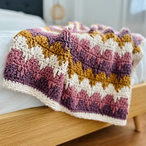 Op de afbeelding: Een gehaakt deken met een chevron-patroon in tinten roze, paars, crème en mosterdgeel. Het deken is gedrapeerd over een houten bedframe.
