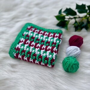 May include: A crocheted blanket with a geometric pattern in green, white, and burgundy. Three balls of yarn in the same colors are in the background.