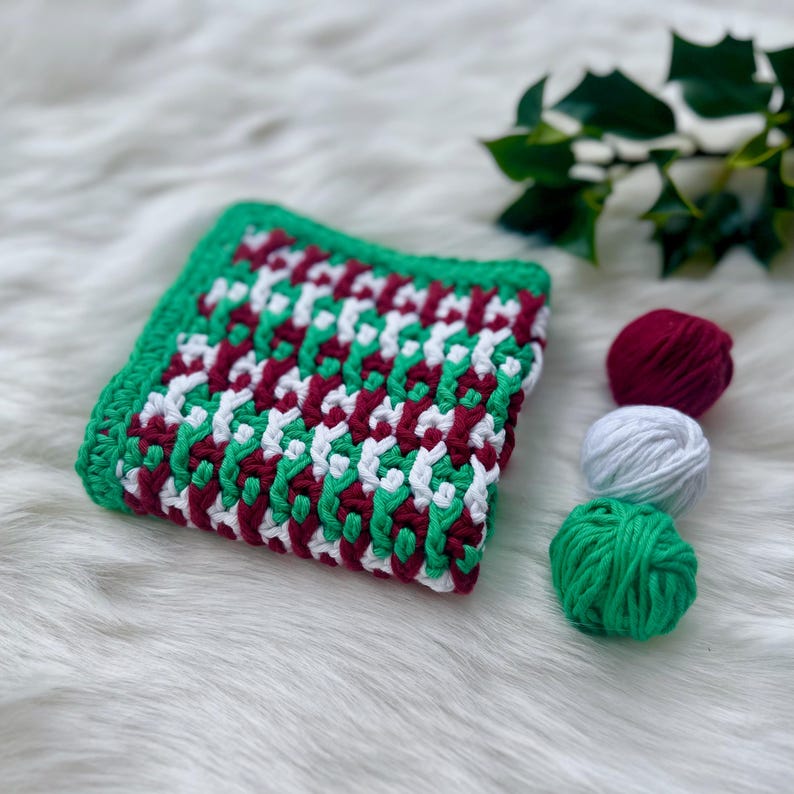May include: A crocheted blanket with a geometric pattern in green, white, and burgundy. Three balls of yarn in the same colors are in the background.