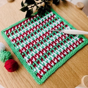 May include: A green, white, and red crocheted square dishcloth with a crocheted border. The dishcloth is lying on a wooden surface with a crochet hook and balls of yarn.
