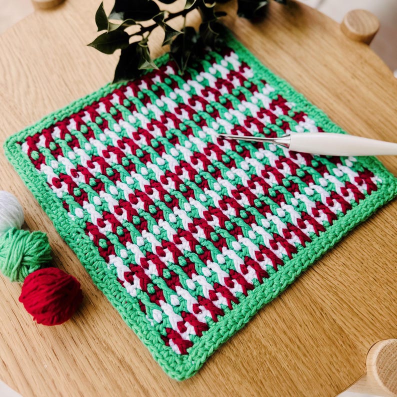 May include: A green, white, and red crocheted square dishcloth with a crocheted border. The dishcloth is lying on a wooden surface with a crochet hook and balls of yarn.