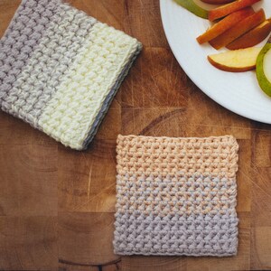 May include: Two crocheted trivets, one in beige, gray, and peach, and the other in white and gray. The trivets are on a wooden surface.