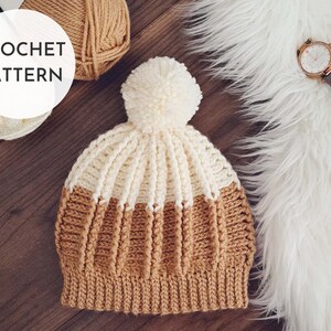 May include: A crocheted beanie hat with a white pom pom on top. The hat is made with two colors of yarn, brown and white, and has a striped pattern. The hat is lying on a wooden surface with a ball of yarn and a watch in the background.