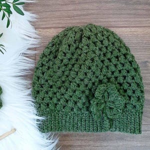 May include: A green crocheted beanie with a flower detail. The beanie is made with a textured stitch and has a folded brim.