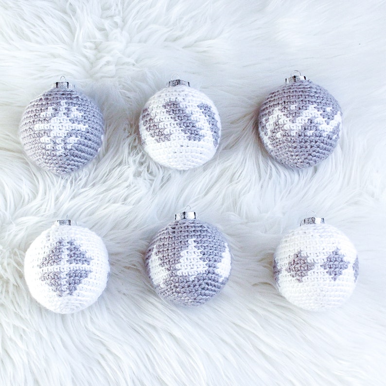 May include: Six spherical ornaments featuring crochet covers in white and grey. Each ornament displays a unique pattern, including snowflakes, geometric shapes, and pine trees, topped with a silver metal cap for hanging purposes.