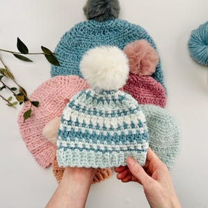 May include: A collection of handmade crochet hats in various colors, including blue, pink, and tan. Each hat features a fluffy pom-pom on top. The hats are displayed on a white surface, showcasing the different textures and designs.