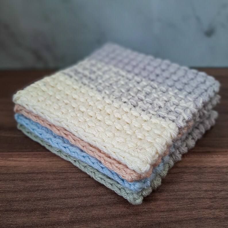 May include: A stack of four crocheted squares in shades of light blue, pink, and white. The squares are made with a textured stitch and are folded neatly.