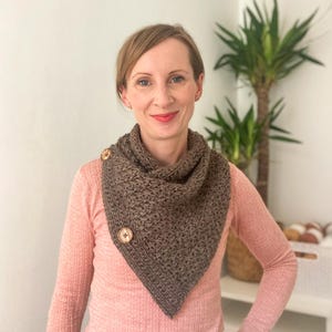 May include: A brown crocheted scarf with two wooden buttons. The scarf is triangular in shape and appears to be handmade. The woman in the image is wearing a pink long-sleeved shirt.