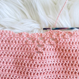 May include: Close-up of a pink crocheted fabric with a delicate openwork pattern. The fabric is being worked on with a crochet hook and yarn.