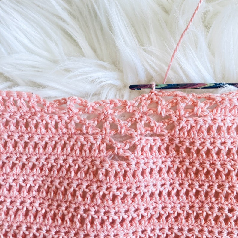May include: Close-up of a pink crocheted fabric with a delicate openwork pattern. The fabric is being worked on with a crochet hook and yarn.