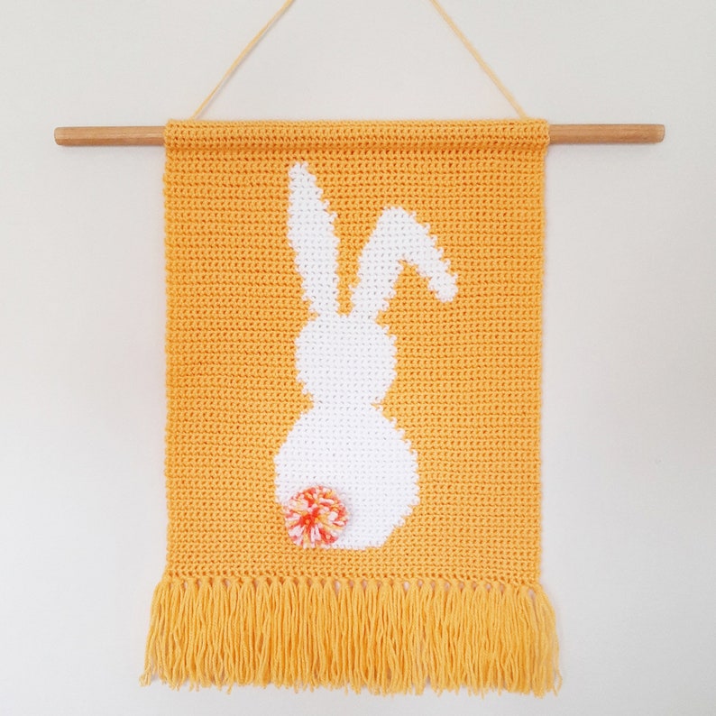 May include: A yellow crocheted wall hanging with a white bunny silhouette and a small pom-pom flower. The hanging has a fringe border.