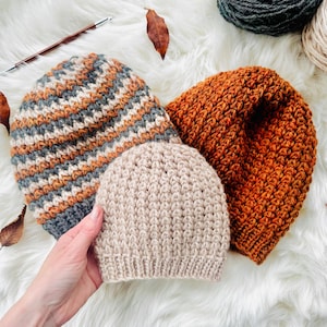 May include: Three handmade knit beanies in shades of orange, gray, and beige. The beanies are arranged on a white, fluffy surface. The largest beanie has a textured, orange pattern. The smallest beanie is beige and has a ribbed texture.