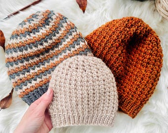 May include: Three handmade knit beanies in shades of orange, gray, and beige. The beanies are arranged on a white, fluffy surface. The largest beanie has a textured, orange pattern. The smallest beanie is beige and has a ribbed texture.