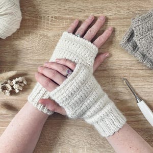 Könnte beinhalten: Cremefarbene, gehäkelte Fingerhandschuhe. Die Handschuhe sind aus dickem Garn gefertigt und haben eine gerippte Manschette. Ein Häkelhaken und ein Wollknäuel sind ebenfalls sichtbar. Die Handschuhe werden auf einer Holzoberfläche präsentiert.