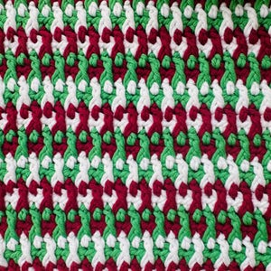 May include: A close-up of a knitted fabric with a repeating pattern of red, white, and green yarn. The fabric has a diagonal weave.
