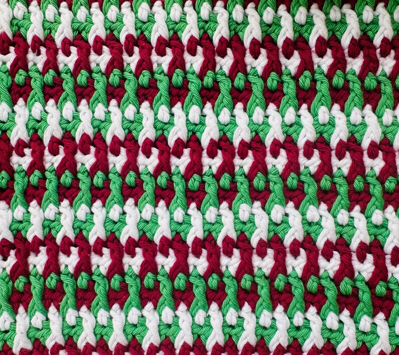 May include: A close-up of a knitted fabric with a repeating pattern of red, white, and green yarn. The fabric has a diagonal weave.