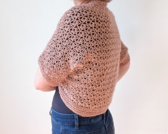 May include: A crocheted bolero in a light brown color. The bolero has short sleeves and a cropped length. It is made with a delicate stitch pattern.