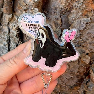 Può includere: Un porta badge con un'immagine a cartone animato del personaggio Ghostface del film Scream. Il personaggio è sdraiato su una nuvola rosa con i piedi rosa. Il porta badge ha il testo "So... What's your favorite scary movie?"