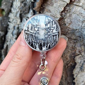 May include: A black and white illustrated badge reel featuring a scene from Diagon Alley in the Harry Potter series. The reel is made of clear plastic and has a silver metal clip.