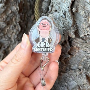 Puede incluir: Un carrete retráctil de insignia redondo y transparente con una imagen de dibujos animados de una persona con el texto "CPR CERTIFIED". El carrete tiene un clip de metal plateado y una correa de plástico transparente.