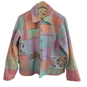 May include: A multi-colored patchwork jacket with a button-up front and two pockets. The jacket features pastel shades of pink, orange, green, and blue. The jacket has embroidered floral details and is hanging on a wooden hanger.