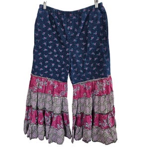 May include: Pull-on pants featuring a dark blue denim upper section with a small pink floral print and a wide-leg silhouette. The lower portion consists of three tiered, ruffled layers in alternating magenta and grey floral patterns.