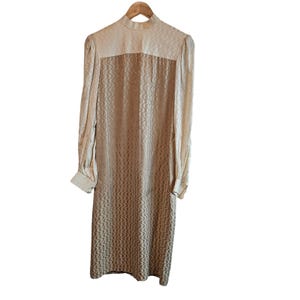 May include: A long-sleeved dress featuring a high neckline and a two-tone color scheme. The upper portion is a light cream, while the main body displays a darker tan hue. Both sections are covered in a repeating, vertical wavy pattern.