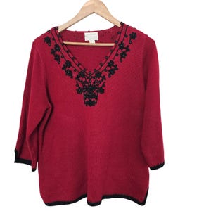 May include: A red long-sleeved sweater featuring a V-neckline adorned with black floral embroidery and beadwork. The sweater includes black trim along the cuffs and the bottom hem, with a label at the inner neckline reading Oscar de la Renta.