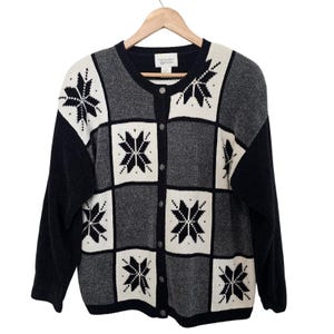 May include: A long-sleeve button-up cardigan featuring a black, white, and gray checkered pattern with black snowflake motifs in each square. The garment has a round neckline, a front button closure, and a Christopher & Banks brand label.