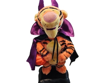 May include: A plush Tigger toy dressed in a Halloween costume. The orange and black striped tiger wears a purple cape and devil horns. He holds a small orange pumpkin-shaped bag. The toy stands on a black base.