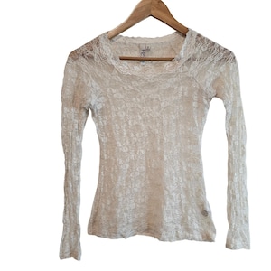 May include: Off-white, long-sleeved lace top. The top is made of a delicate floral lace fabric and features a scoop neckline. The top is hanging on a wooden hanger.