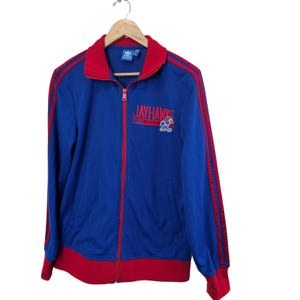 May include: Blue zip-up track jacket with red accents. The jacket features red stripes down the sleeves and red trim at the collar, cuffs, and hem. The word "JAYHAWKS" is embroidered on the chest, along with a mascot.