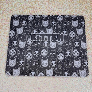 May include: Black rectangular mouse mat featuring a repeating pattern of cat faces, skulls, and pentagrams. The word "KITTEN" is in white, bold letters in the centre. The background is a glittery, iridescent surface.