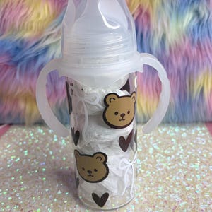 May include: A clear baby bottle with a white nipple and handles. The bottle is decorated with brown bear faces and dark brown hearts. The bottle is filled with white paper and is on a glittery surface.