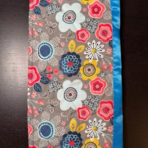 Puede incluir: Una manta de bebé acolchada rectangular con un estampado floral en tonos grises, rosas, azules, amarillos y blancos. La manta está ribeteada con un borde de satén azul brillante. El diseño floral incluye flores grandes y pequeñas, mariposas y hojas.