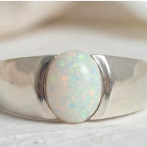 Puede incluir: Un anillo de plata con una gran piedra de ópalo ovalada. El ópalo tiene un color base blanco lechoso con destellos de colores iridiscentes. La banda del anillo es ancha y lisa, con un diseño simple y moderno.